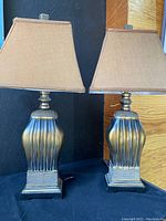 Pair of two table lamps with sculpted metallic bases and brown rectangular double lined shades shown side by side against dark background and plywood panel.