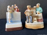 Front view of two music box figurines on black surface. Left figurine shows a nurse and older couple by hospital counter, right figurine shows older couple by stove.