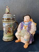 Photo showing 10 Staffel West Germany stoneware beer stein with detailed painted scenes and decorative metal lid topped with a small figure, alongside Sunley seated male musical figurine.