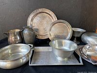 Seven metal items displayed on black surface: two round pewter trays, a rectangular hammered aluminum tray, a silver on copper tray, a pewter footed bowl, a pewter vase, and a metal pitcher.