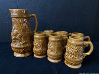 Set of one large ceramic pitcher and six matching beer steins, all brown glazed with relief designs, shown together against dark background