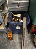 Blue bin with various hand tools and vintage lights inside, orange 12 ton hydraulic bottle jack on floor next to it, heavy metal bar with handle leaned against bin