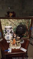 Photo showing entire lot on a wooden table: capodimonte vase, framed print in background, black metal lantern, and ceramic figurine.
