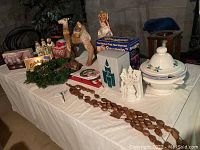 Table showcasing a hand-painted soup tureen with floral motif, wooden rosary beads stretched across, holiday decorations including Christmas wreath, Three Kings figurines, camel, Christmas tree topper, votive candle holders and a box of Snuggle.
