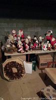 Wide shot of a table with about 16 Santa Claus figurines in various outfits and poses, some with accessories, set against a brick wall. Below the table is a pine cone wreath.