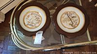 Two Westland wood framed collector plates with Japanese-style bird and blossom illustrations displayed on a glass table.
