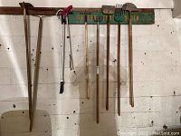 Photo of 9 hanging garden tools on a wall; includes rakes, edgers, hoe, cultivator, and long stick