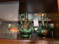 Shelf display showing two green glass ewers and six matching stemware glasses with gold floral hand painted designs.
