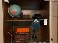 Shelf with two men's hats, terrestrial globe, wooden chest with glass decanter and shot glasses, and green embroidered tie.