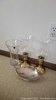 Two brass hurricane lamps with clear glass chimneys, one clear glass vase and one Caravelle silver plate tray with handle on carpeted floor against wall.