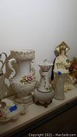 Photo showing large ornate porcelain vase with floral and scroll detailing, smaller vases and religious figurine on shelf.