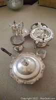 Overall view of silver plated serving set including coffee pitcher on warming stand, multiple serving bowls and trays with lids featuring ornate details and some tarnish.