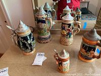 Five decorative beer steins with pewter lids displayed together showing variety of designs and colors.