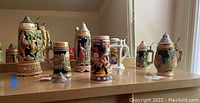 Seven beer steins arranged on wooden table with natural light from window. Varied sizes and traditional German and sports themes.