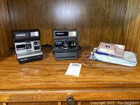 Full view of the three cameras displayed on wooden surface: Polaroid Sun 600 LMS, Polaroid OneStep Closeup, and LeClic digital camera with bag.