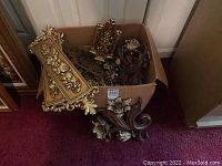 The lot items inside a cardboard box with several gilded and dark finish ornate wall accents placed upright and overlapping each other. Mainly showing the top edges and some details of the gilded flower and scroll carvings.