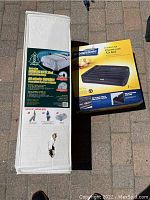 Photo showing two boxed inflatable beds: Woods Twin Size BYO Bed with built-in pump on left, Broadstone Double High Queen Size Air Bed on right, outdoors on pavement.