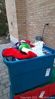 Blue plastic bin filled with Christmas decorations and boxes, placed outdoors on paved ground near brick building.
