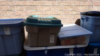 Stacked plastic storage bins containing Christmas decorations and a box of crystal glasses on a daylight brick background.