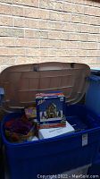 Large blue storage bin with lid open showing packed items including boxed village buildings and musical snowglobe box.
