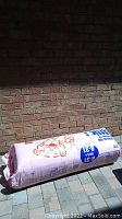 Photo of full roll of pink fiberglass insulation still in packaging resting against a brick wall outdoors.