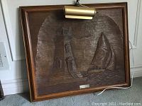 Frontal view of the framed wood carving artwork showing the lighthouse and sailboat scene with mounted light fixture on the top edge.