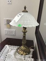 Front view of table lamp on lace doily on wooden surface, showing brass base and frosted glass shade with scalloped edge.