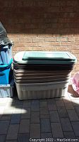 Stack of five beige Rubbermaid plastic storage totes with green lids, stacked outdoors against a brick wall.