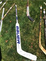 Overview photo of the white Bauer Supreme 405 goalie stick with blue branding lying on grass, showing multiple hockey sticks, one prominently in the center with space for signature.