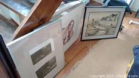 Photo of three framed art pieces leaning against a low wall and an angled wooden beam on a wooden floor, showing relative sizes and styles of artworks.