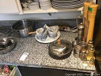 Multiple stainless steel mixing bowls stacked, two marble mortar and pestle sets on lazy Susan, metal and wood salt and pepper grinders and shakers, and other kitchenware on countertop.