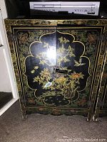 Front view of the black painted cabinet showing floral and bird designs and metal latch.