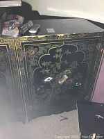 Front view of the black lacquer cabinet showing floral and bird hand-painted artwork and scratches on the top surface.