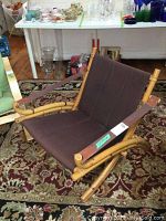 Front-right angled view of wood frame chair with brown canvas seat on a decorative rug, background shows various household items