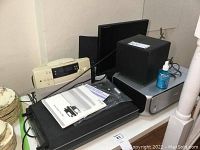 Multiple electronic items on a table including Epson scanner, Canon printer, Aztec Lansing speakers, Princeton monitor, UBL player, and some manuals and small bottle of cleaner