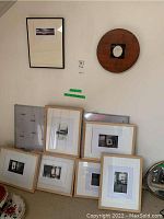 Six framed black and white photos resting on a carpeted floor under an angled ceiling. Two pieces of art visible on the wall above, one circular wood piece with photo and one framed photo. Frames are natural wood Ikea frames measuring 13x16 inches.