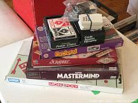 Stack of board games on a white table including Parcheesi, Scrabble, Mastermind, Monopoly, with poker chips and card decks on top