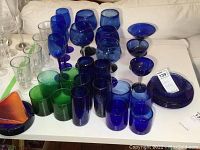 Overview of various wine glasses, tumblers, in blue, green, and clear glass placed on white surface.