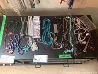 Top view of assorted costume jewelry necklaces laid out on a black table surface, showing various styles and colors including white faux pearl, turquoise beads, metal chains, and others.