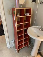 Front view of a tall laminate shelf unit with 14 square cubbyholes. Shelf is positioned beside a pedestal sink and door frame. Small items displayed inside cubbies are not included in sale.