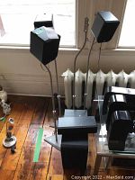 Overview of several black speakers mounted on silver metal stands with triangular bases, placed in front of a window and radiator, showing their size and design.