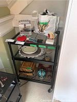 General view of the lot items including boxes, bowls, tray, vintage sterling hairbrush, jewelry vases, plastic lockbox, bag of hair bands, and a white tote bag on a black shelving unit.