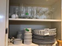 Shelf with stacked white ceramic dishware including pie plates, ramekins, large plates. Above shelf shows clear glasses including tall glasses and martini glasses with stems.