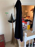 Black velvet witches cape draped over broom and witch hat standing against wall.