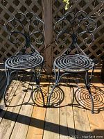Front view of a pair of black aluminum bistro chairs with round spiral seats and decorative scrollwork backs on a wooden deck.