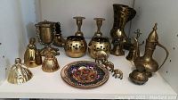 Full shelf view showing all 18 brass items including lidded pot, candleholders, boots, bells, plate, and figurines