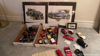 Photo showing two framed vintage car art prints behind boxes containing about two dozen collectible model cars in multiple colors including red, yellow, white, blue, and black.