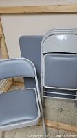 Photo showing grey metal folding card table top alongside three folded chairs leaning against the wall, highlighting the grey vinyl padded surfaces and metal frames