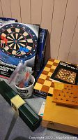 Photo showing Unicorn electronic dartboard with blue darts, Halex bristle dartboard, chess board, and a wooden box likely holding chess pieces or a game.