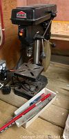 Compact black and metal Jobmate drill press with drill bits in a small box in front of it, showing the overall look and accessories.